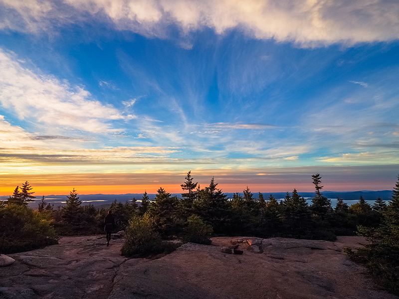 Photo of single person walking toward sunrise at top of mountain in Acadia National Park.