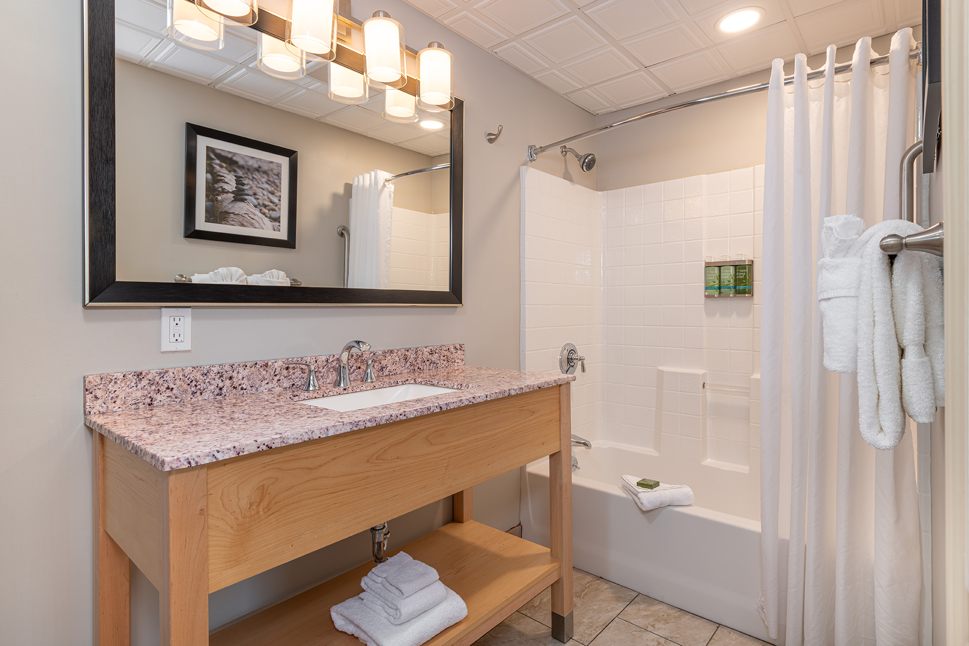 A clean bathroom with a granite countertop sink, light wood furniture, and a tiled shower and tub.